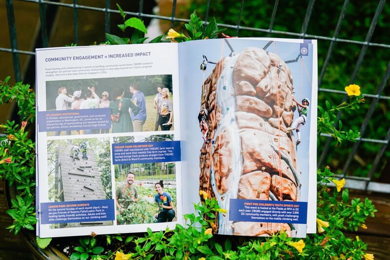 The image shows an open magazine with a focus on community engagement and impact. The left page features a collage of photos depicting people involved in community activities, such as gardening and climbing. The right page displays a large, abstract sculpture or art installation. The magazine is surrounded by greenery, suggesting a connection to nature or outdoor community projects.